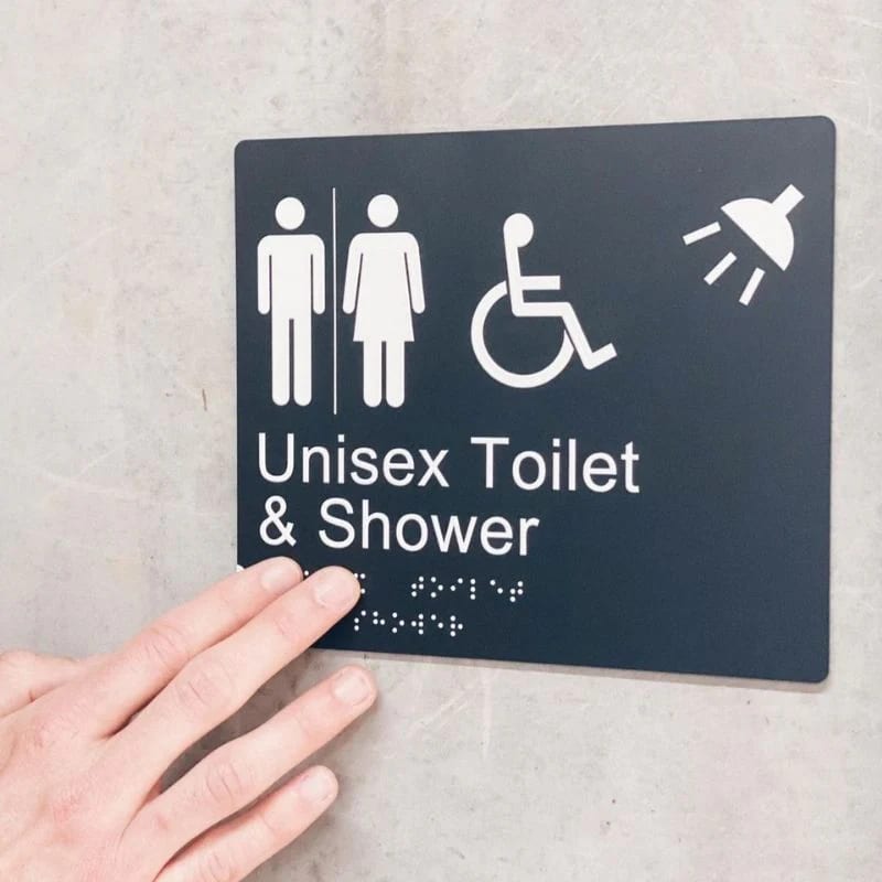 A sign reads "Unisex toilet and shower". Beneath the visual text is braille writing. A person's hand is touching the braille in order to read it.