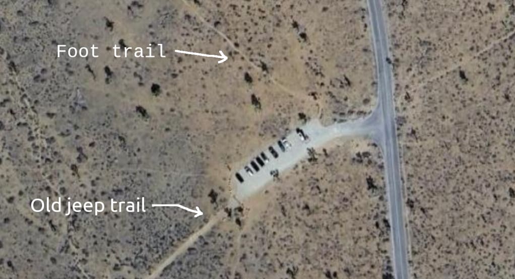 Satellite view of parking lot off a road in the wilderness. Out of the parking lot, from the air, we see one faint curving foot trail, and on the other side of the lot, one very clear wide jeep trail.