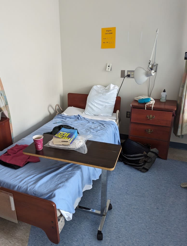 My hospital bed with folded scrubs atop, a cup of coffee, and copy of Infinite Jest set on the adjustable bedside table.