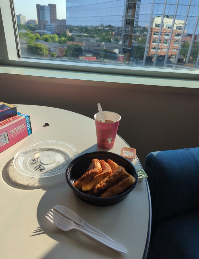 Breakfast - french toast in a plastic container and a cup of coffee - illuminated in golden morning light, at a table with a nice view out into the city.